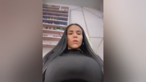 Video of a woman with long, straight black hair, wearing a tight black top, standing in a brightly lit room with shelves of colorful nail polish bottles in the background.