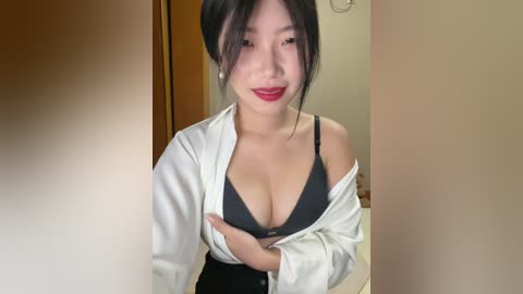 Video of an Asian woman with fair skin, black hair, and red lipstick. She wears a black bra, white shirt, and black pants, posing indoors with a wooden door and beige walls in the background.