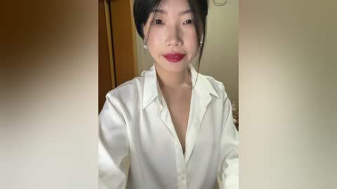 Video of a young Asian woman with fair skin, black hair tied back, wearing a white silk shirt, smiling softly, indoors with wooden door and beige wall background.