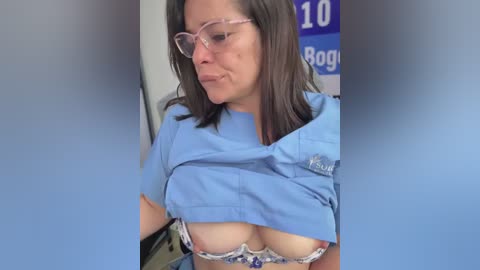 A video of a Caucasian woman with glasses, wearing blue scrubs, lifting her top to reveal a floral bra. She has medium-sized breasts and dark hair. The background shows a hospital sign with \"10 Rooms\" and \"Bogus.\