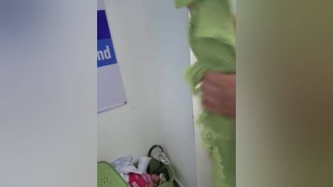A video shows a person in a green uniform, possibly a nurse, standing in a dimly lit room with a white wall and a blue \"NHI\" sign. A green laundry basket with clothes is on the floor.