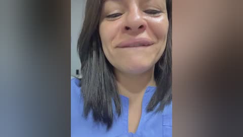 Video of a smiling woman with medium skin tone, shoulder-length dark hair, wearing a light blue V-neck shirt. The background is blurred, emphasizing her face.