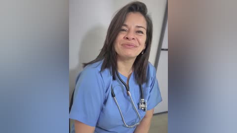 Video of a smiling, middle-aged woman with light skin and shoulder-length dark hair, wearing light blue scrubs and a stethoscope, standing in a hospital hallway.