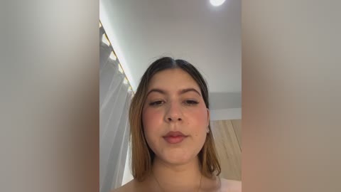 Video of a young Latina woman with light brown skin, straight brown hair, and brown eyes. She wears a soft pink top, standing in a modern, well-lit room with beige walls and a wooden cabinet.