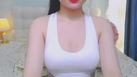 A video of a fair-skinned Asian woman with long black hair, wearing a white tank top, sitting on a bed with a beige quilt. The room has a nightstand with a lamp, and the background features a soft, pastel-colored wall.