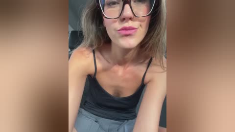A close-up video of a fair-skinned woman with straight, shoulder-length blonde hair, wearing black-framed glasses and a black spaghetti-strap top, sitting indoors with a neutral background.