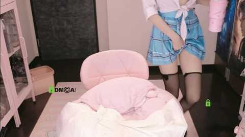 A video shows a light-skinned woman in a blue plaid skirt and white top, standing near a pink chair in a room with a white bookshelf, pink blanket, and green \"DMGAI\" watermark.