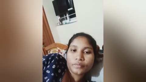 Video of a young woman with medium skin tone, long black hair, and a sad expression, lying on a bed with a blue patterned blanket. Background features a wooden chair, white wall, and a black TV.