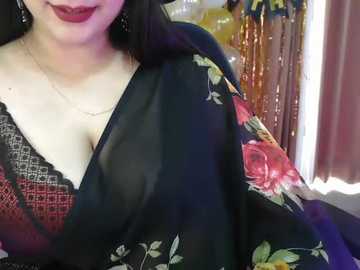 Video of a woman with fair skin, long black hair, and red lipstick, wearing a black floral saree with a red lace bralette underneath. Background features a golden curtain and a blurred, colorful room.