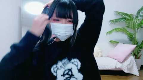 Video of an Asian woman with straight black hair, wearing a black sweater featuring a bear graphic, a face mask, and adjusting her glasses in a modern, minimalist room with a green plant and white sofa.