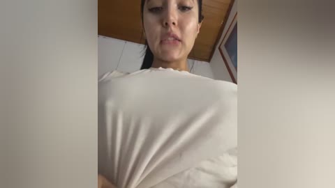 A video of a woman with light skin, dark hair, and a neutral expression, wearing a white blouse that reveals her cleavage. The background features a wooden ceiling and part of a framed picture.