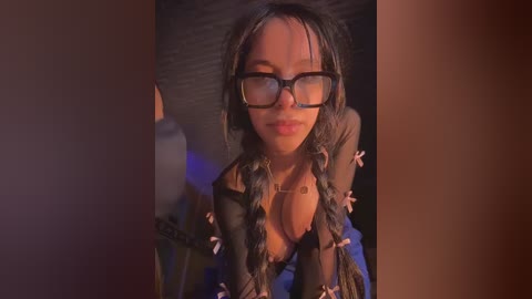 Video of a young woman with light brown skin, wearing black-framed glasses, braided hair, and a low-cut, black, sequined top, leaning forward, in a dimly lit room with a dark background.