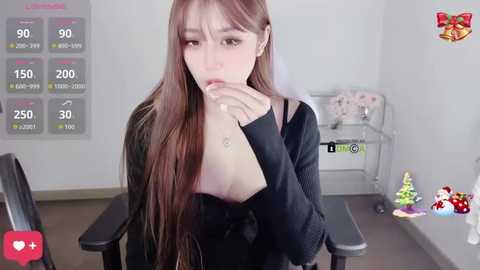 A video of an Asian woman with long, straight brown hair and pale skin, wearing a black top, looking pensively at a smartphone in a sterile room with a medical cart and temperature gauge.