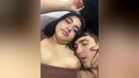 Video of a young, light-skinned woman with medium-length black hair and a small bust, lying on her back. A young man with short brown hair and medium skin tone, wearing a brown shirt, lies next to her, kissing her neck.