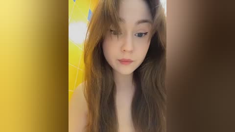 A video of a young, fair-skinned woman with long, wavy brown hair, wearing minimal makeup, standing in a yellow-tiled bathroom, with a blurred background.