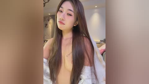 Video of an Asian woman with long, straight brown hair, wearing a white robe, partially exposing her small breasts. She has light skin and a delicate facial structure. The background is a softly lit bedroom with neutral tones.