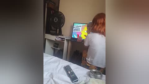 Video of a young woman with red hair, wearing a white T-shirt, sitting on a bed, watching TV on a white dresser.