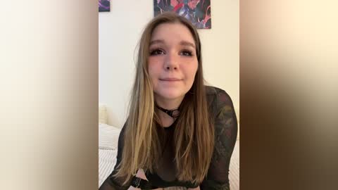 A video of a young Caucasian woman with long, straight, light brown hair, wearing a black sheer top, smiling, standing in a softly lit bedroom with a white bed and colorful abstract art on the wall.