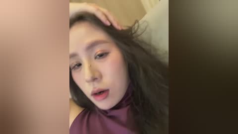 Video of an Asian woman with long, dark hair, light skin, and a high ponytail, wearing a purple top, looking contemplative with her hand on her head.