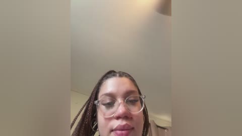 A video of a young woman with medium brown skin, wearing glasses and braided hair, taken from a low angle. She has a neutral expression and is indoors against a beige wall.