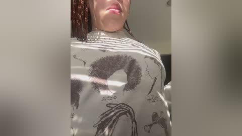 Video of a woman in a gray shirt featuring anatomical drawings. Her lips are slightly parted, and she has shoulder-length, curly brown hair. The background is blurred, showing a white wall and part of a door.