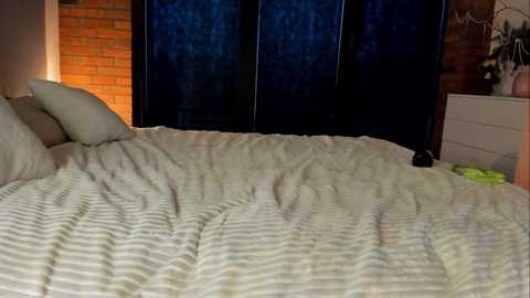 Video of a dimly lit bedroom with a white striped bedspread, a white pillow, brick wall, and dark blue tinted windows.