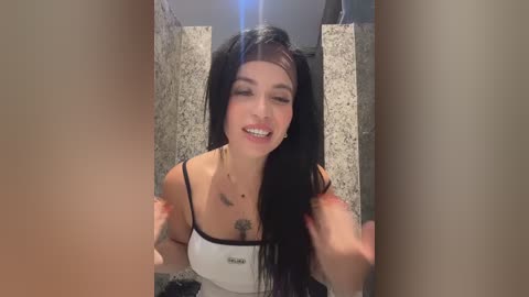 Video of a smiling, slender, light-skinned woman with long black hair, wearing a white tank top and black bra, standing in a bathroom with a speckled granite countertop.