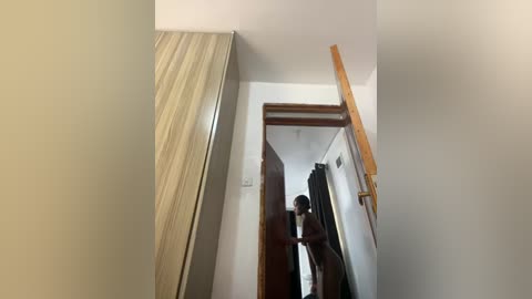 Video of a man with a dark complexion and short hair, standing naked in a dimly lit hallway with beige walls and a wooden door.