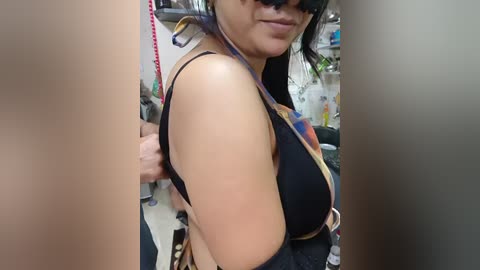 Video of a woman with light skin and black hair, wearing a black bra and a colorful, patterned bag, standing in a cluttered room with shelves and bottles in the background.