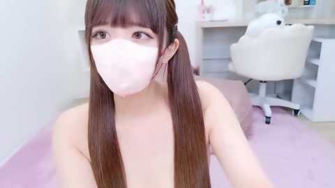 A video of a young Asian woman with long, straight brown hair, wearing a white surgical mask, seated on a pink bed in a minimalist, bright room with a white desk and chair.