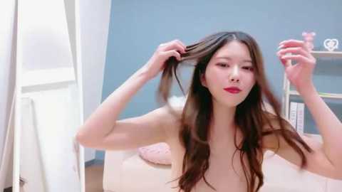 A video of an Asian woman with long brown hair, red lipstick, and light skin, combing her hair in a modern, minimalist bedroom with blue walls and a white bed.