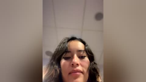 Video of a young woman with wavy dark hair, fair skin, and light makeup, looking down, indoors with a beige tiled ceiling and a blurred background.