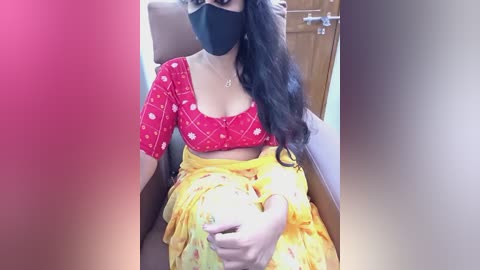 Media: A video of a South Asian woman with medium skin tone, long black hair, and a red mask, wearing a red top with white patterns and a yellow sari with floral print, seated in a brown chair.