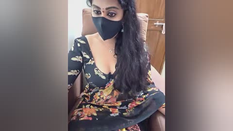 A video of a South Asian woman with long, wavy black hair, wearing a floral dress, black face mask, and seated on a brown leather chair in a well-lit room.