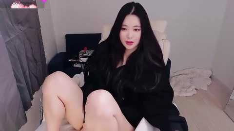 A video of an East Asian woman with long black hair, fair skin, and red lipstick, wearing a black jacket, sitting on a bed with white sheets. Background features a gray curtain, a black chair, and a cluttered nightstand.