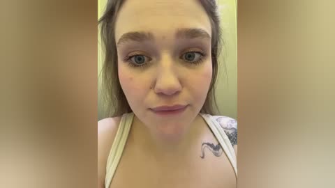 Video of a young woman with fair skin, blue eyes, and light brown hair. She wears a white tank top, revealing a tattoo on her right shoulder. The background is blurred, focusing on her face.