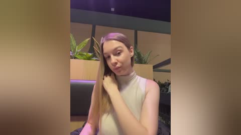 Video of a young Caucasian woman with long, straight blonde hair, wearing a sleeveless white turtleneck. She sits indoors with green plants in the background, smiling and touching her hair.