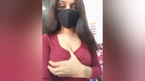 Video of a young woman with medium brown skin, long dark hair, wearing a black face mask, red long-sleeve top, and a necklace. She's indoors with a blurred background.