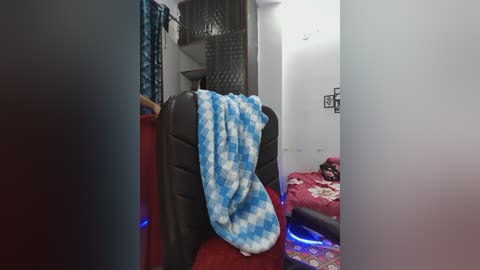 Video of a cluttered bedroom with a blue and white checkered blanket draped over a black chair, red carpet, and a bed with floral bedding in the background.