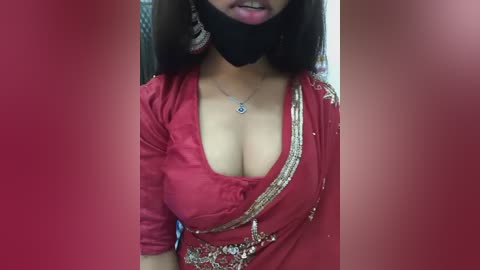 Video of a South Asian woman with medium skin tone wearing a red, gold-embroidered sari, black mask, and earrings, smiling. Background is blurred.