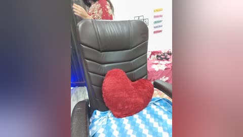 Video of a person with dark skin, seated in a black leather office chair with a red heart pillow. The background shows a bedroom with a white wall and a blue duvet with white polka dots.