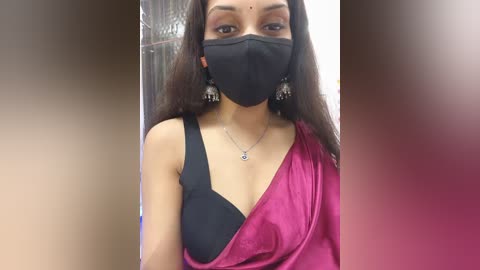 Video of a woman with long black hair, wearing a black mask, black bra, and a pink sari with a black sleeve. She has a silver pendant necklace and ornate earrings. Background is blurred, indoors.
