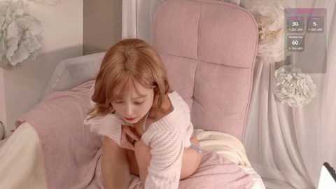 Video of a young woman with light skin and shoulder-length brown hair, wearing a white off-shoulder sweater, sitting on a pink upholstered chair in a soft, pastel-colored room.