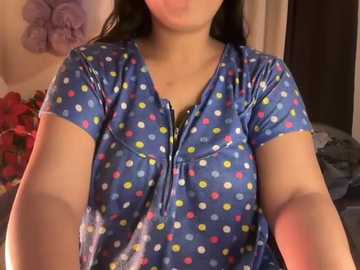 Video of a woman with medium skin tone, dark hair, wearing a blue polka-dotted shirt, posing indoors with a blurred background featuring a purple floral wall and red flowers.