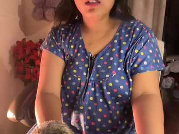 Video of a woman with medium skin tone, wearing a blue, short-sleeved top with colorful polka dots. She's seated indoors, with a vase of red roses and a dark sofa in the background.