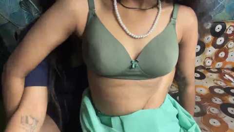 Video of a light-skinned woman with a slim physique wearing a green bra and turquoise skirt, seated on a chair with a patterned background.