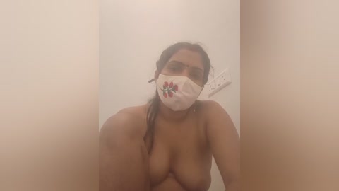 A video of a topless South Asian woman with medium skin tone, wearing a white face mask with a floral design, and brown hair tied back. She stands in a dimly lit, beige-walled bathroom.