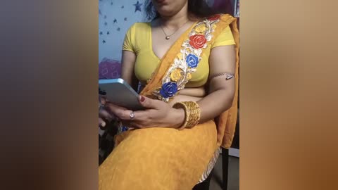 Video of a South Asian woman in a bright yellow sari with a floral border, seated indoors, wearing gold jewelry, holding a smartphone, and smiling.