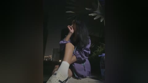 Video of a woman with long black hair, wearing a purple robe and white socks, sitting on a bed in a dimly lit room with a white chandelier and potted plants in the background.
