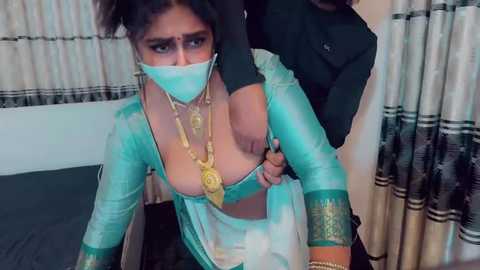 Video of a South Asian woman in a teal salwar kameez, partially exposed, wearing a face mask, held by a man in a black jacket.